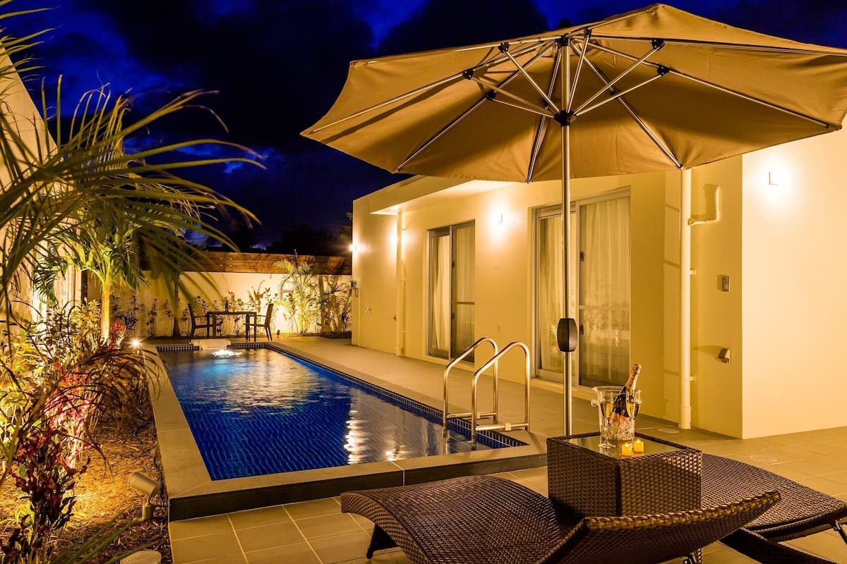A swimming pool with a sun umbrella, lounge chairs, and a well-lit patio area.