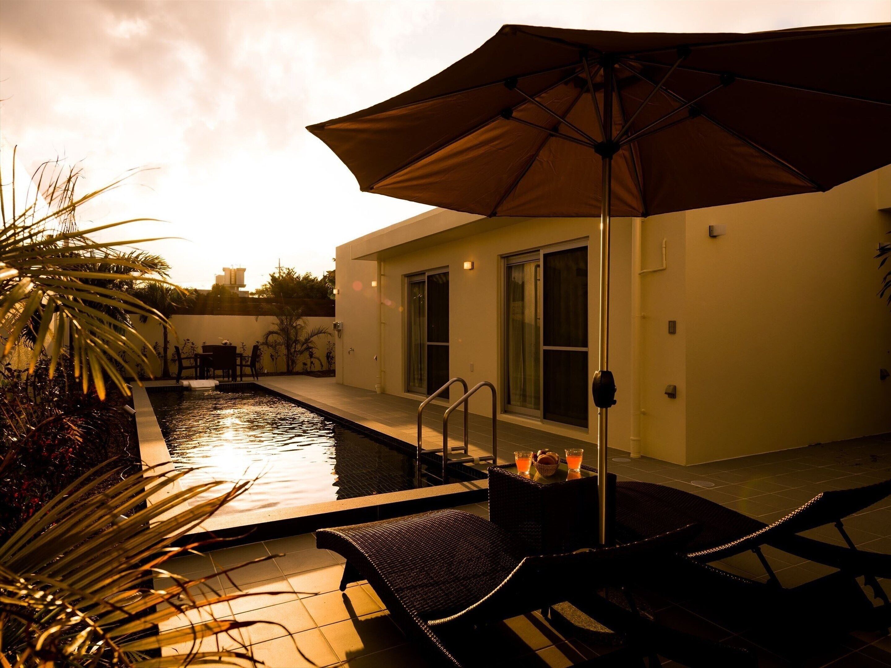 A pool area with a lounge chair and umbrella, surrounded by plants and a building with large windows.
