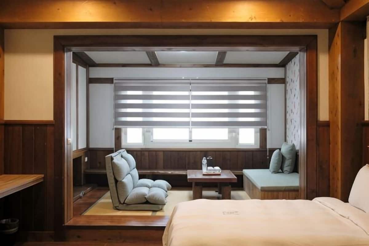 A room with a bed, a chair, a small table, and a window with blinds.
