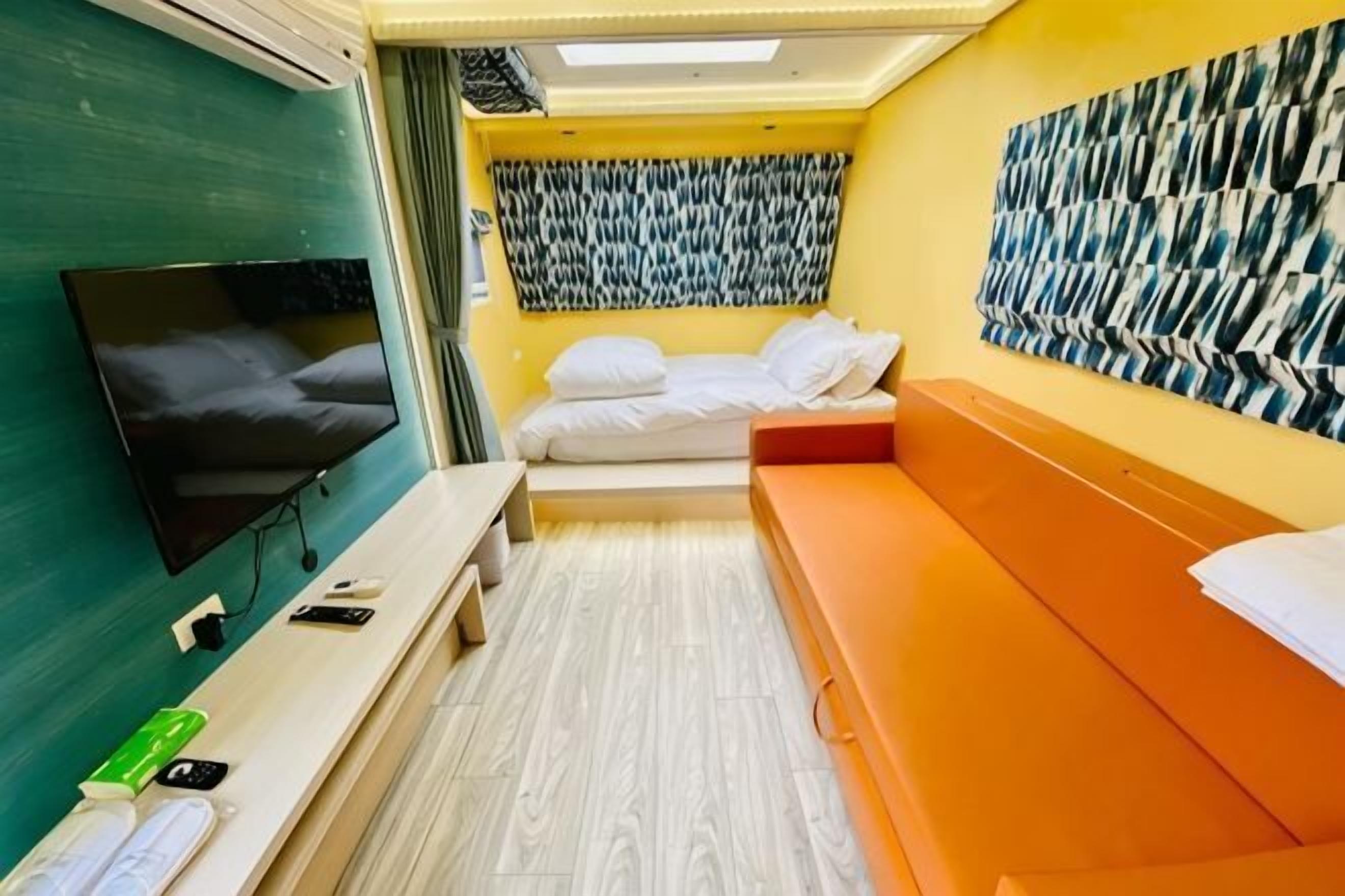 Ein schmaler Raum mit Bett, Sitzbank, Fernseher und Wandbild.