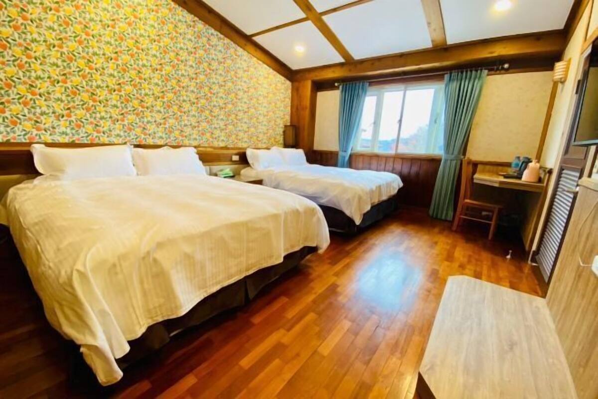 A hotel room with two beds, wooden flooring, a patterned wall, a window with curtains, and a small table with a lamp.