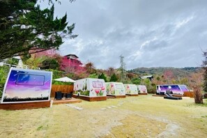 Fileira de cabanas com chão de terra, rodeadas por árvores e uma colina ao fundo.