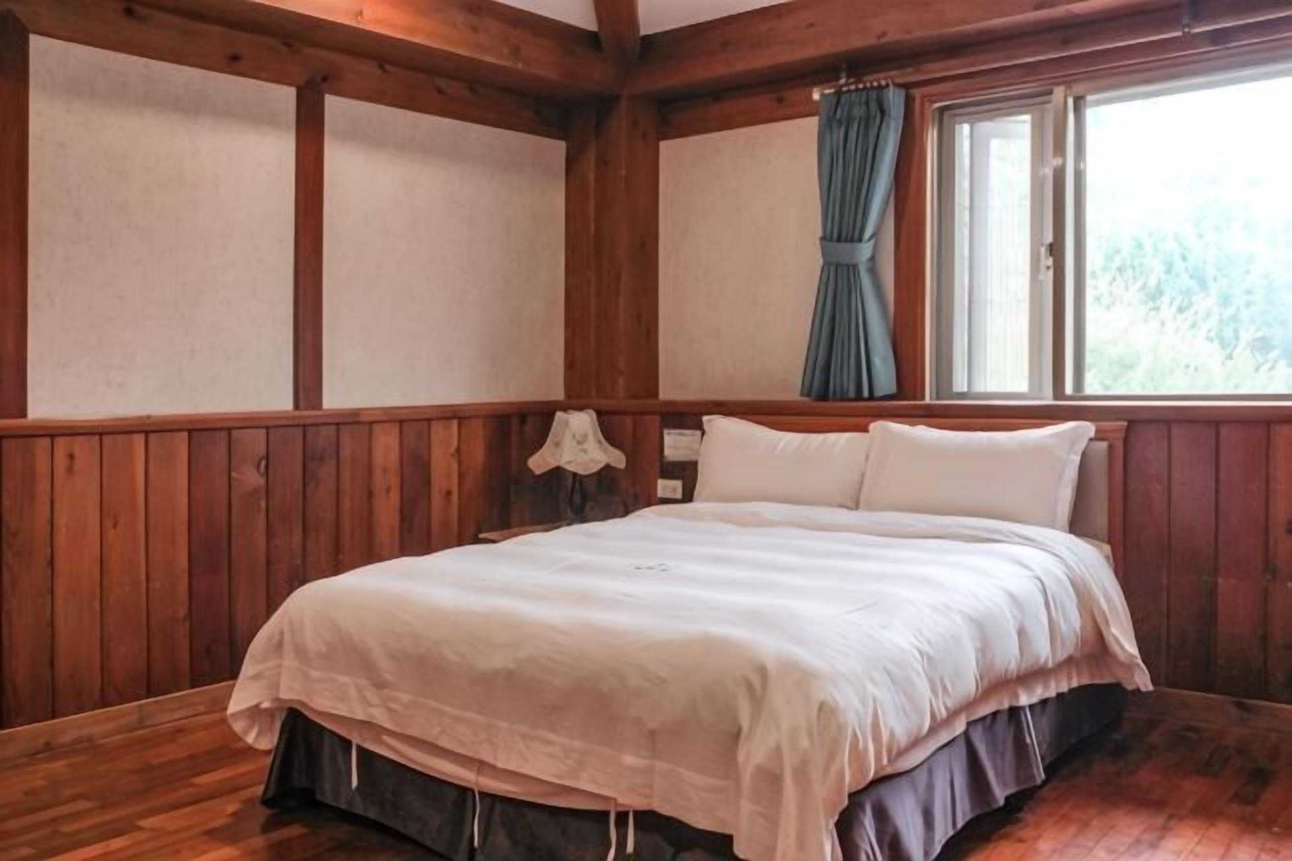 Ein Zimmer mit einer holzverkleideten Wand, einem Bett mit weißer Bettwäsche und einem Fenster mit Vorhängen.