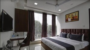 Ein Hotelzimmer mit Bett, Schreibtisch, Stuhl, einem Fenster mit Vorhängen, einem Deckenventilator und einem Bild an der Wand.