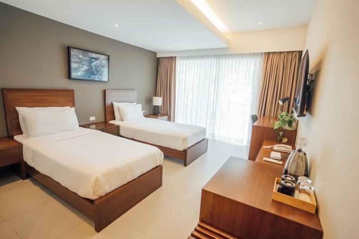 A hotel room with two beds, a desk, a TV, and a window with curtains.