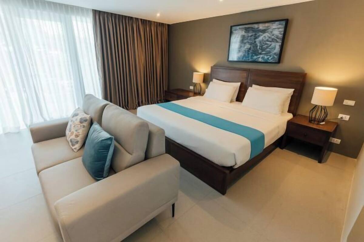 A hotel room with a bed, a sofa, two bedside tables, and a large window with curtains.