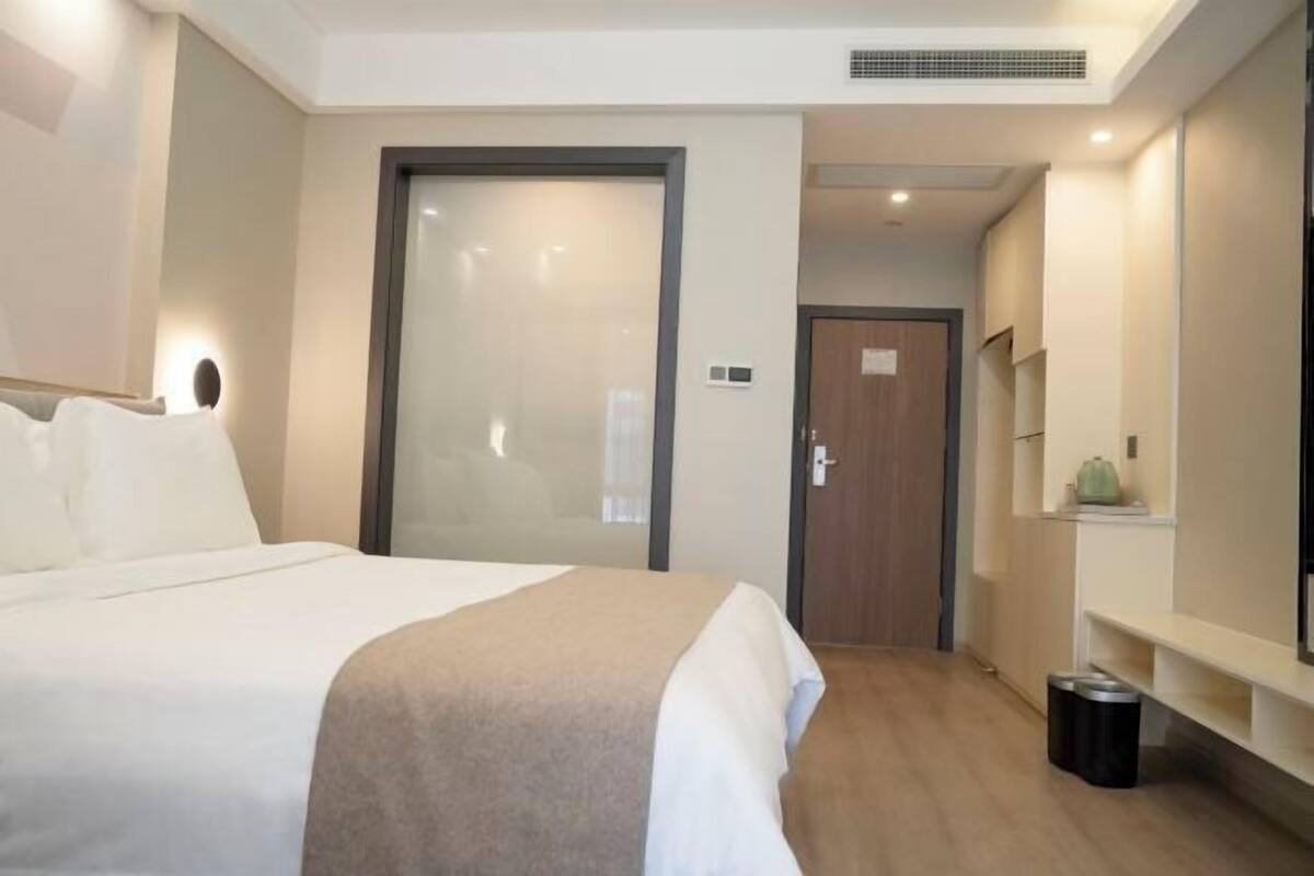 Ein Hotelzimmer mit einem Bett, einer Tür, einer kleinen Mülltonne und einem Regal mit einer Pflanze.