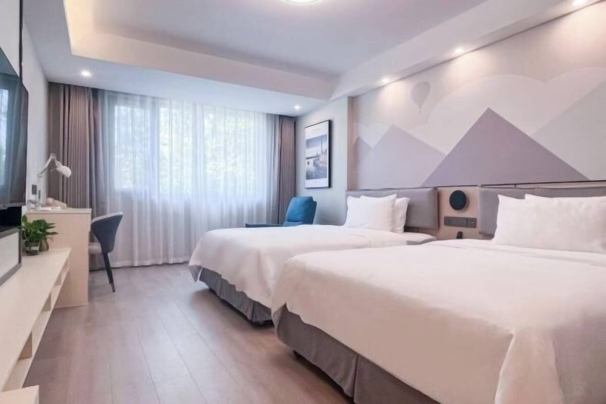 Ein Hotelzimmer mit zwei Betten, einem Schreibtisch, einem Stuhl und einem großen Fenster mit Vorhängen.