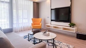 Ein modernes Wohnzimmer mit einer Couch, einem runden Couchtisch, einem leuchtend orangefarbenen Sessel, einem an der Wand befestigten Fernseher und einem großen Fenster mit durchsichtigen Vorhängen.