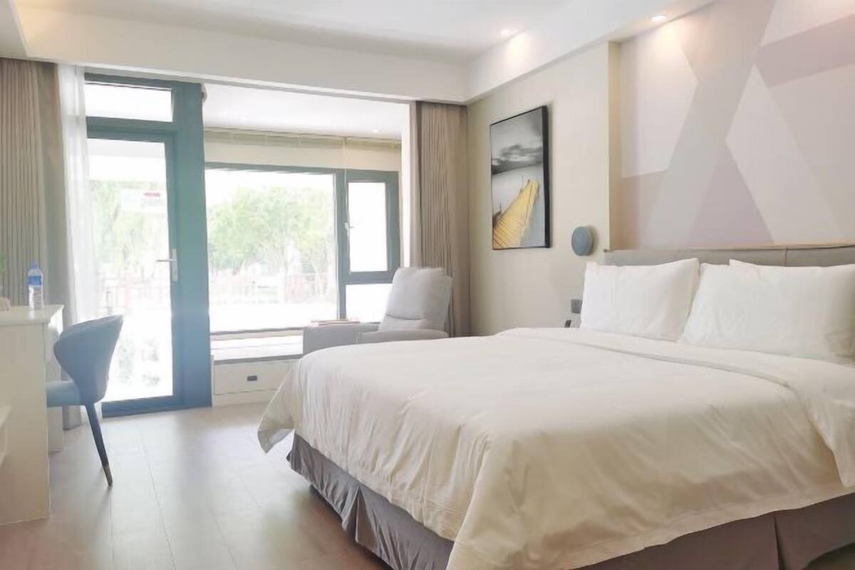Ein Hotelzimmer mit einem großen Bett, einem Schreibtisch und einem Stuhl. Ein Fenster mit Vorhängen bietet Blick auf grüne Landschaft.
