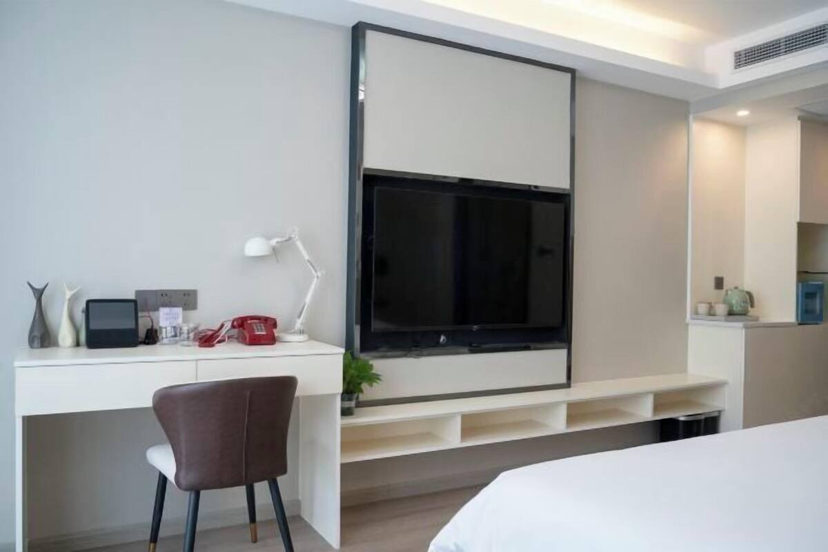 Ein modernes Hotelzimmer mit Wandfernseher, Schreibtisch mit Stuhl, Nachttisch und einer kleinen Küchenzeile.