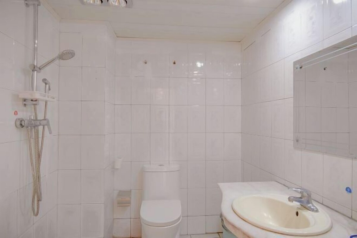 A bathroom with white tiles, a toilet, a shower, and a sink with a mirror.