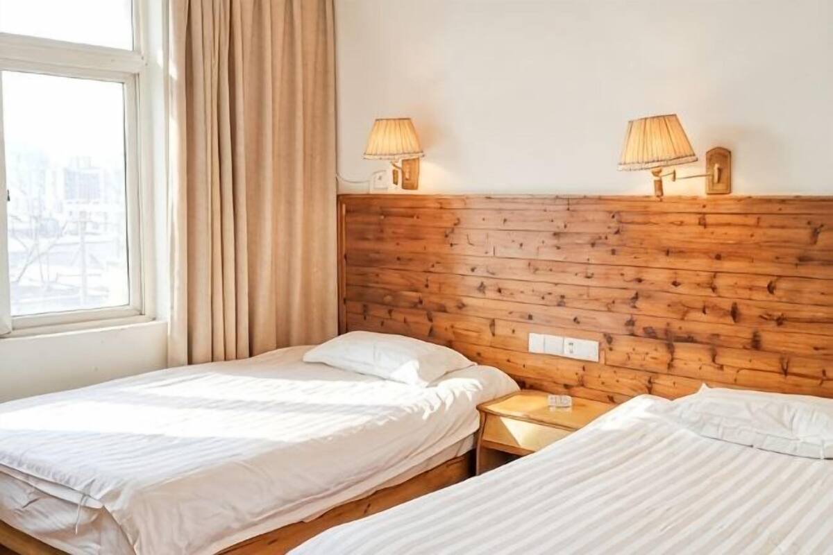 A room with two beds, wooden headboard, bedside lamps, a window with curtains, and a view of the city.