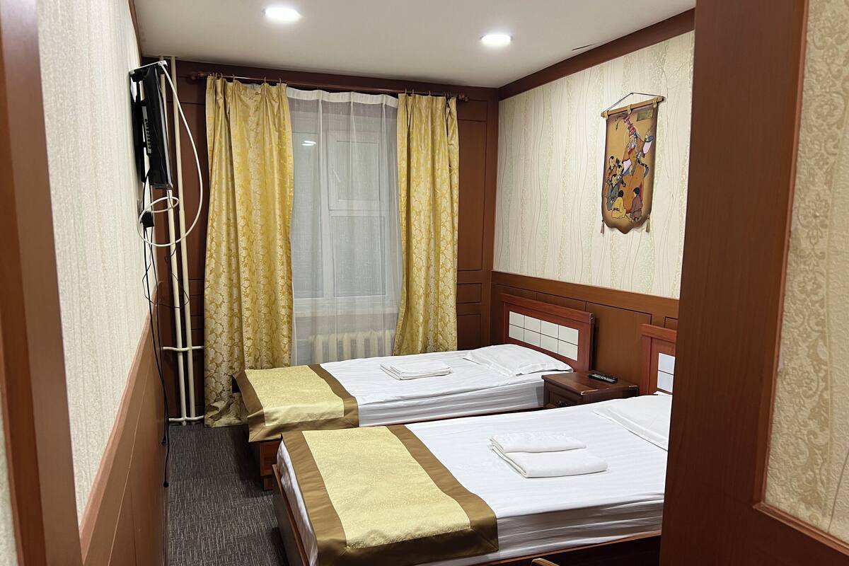 A hotel room with two beds, a wooden headboard, a curtain, a radiator, and a wall-mounted clock.