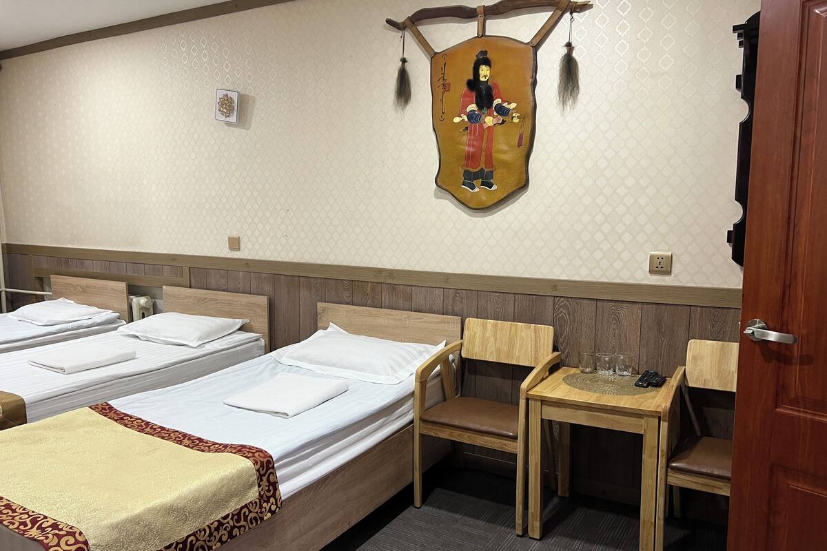 A hotel room with two beds, a wooden headboard, a small wooden table with glasses and a remote control, and a decorative wall hanging.