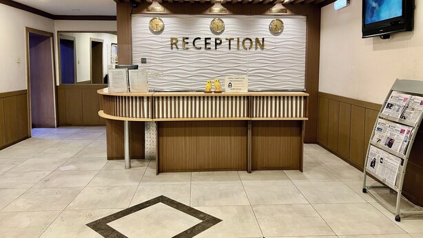 A reception desk with a 'RECEPTION' sign, a flat-screen TV, and a newspaper rack. - Negdelchin Hotel & Service Apartment (Ulaanbaatar)