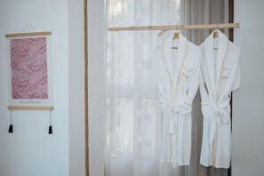 Une pièce dans laquelle on aperçoit deux robes blanches suspendues à un portant en bois, une œuvre d’art encadrée accrochée au mur et une fenêtre ornée de rideaux légers.