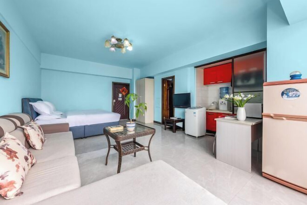 A compact hotel room with a bed, sofa, small table, kitchenette, and a TV.