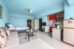 A compact hotel room with a bed, sofa, small table, kitchenette, and a TV.