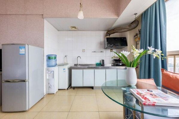 A compact kitchen with a refrigerator, water dispenser, and a sink. There's a glass table with a magazine and a vase with flowers.