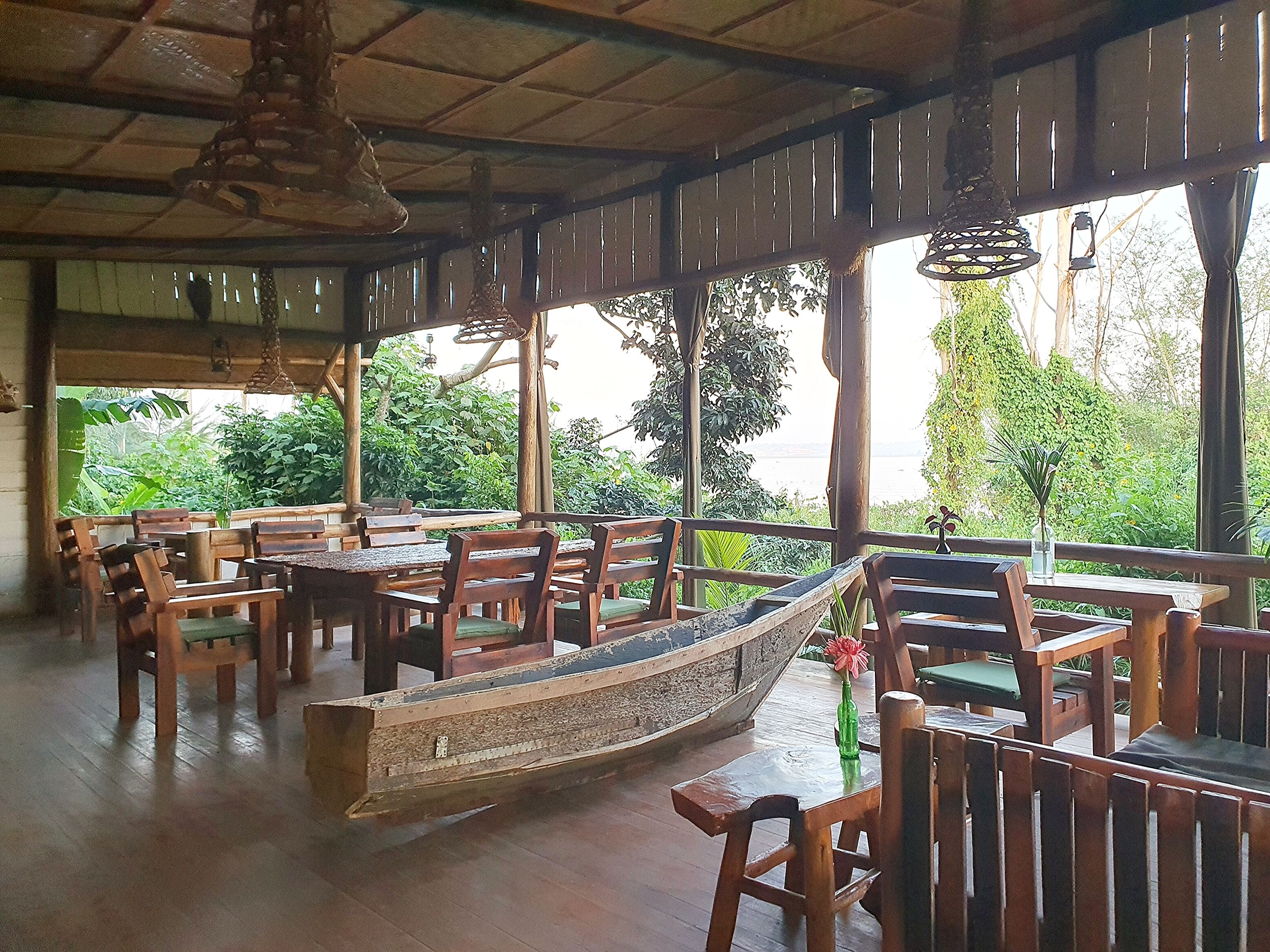 A covered outdoor seating area with wooden tables and chairs, a boat, and a view of greenery and water.