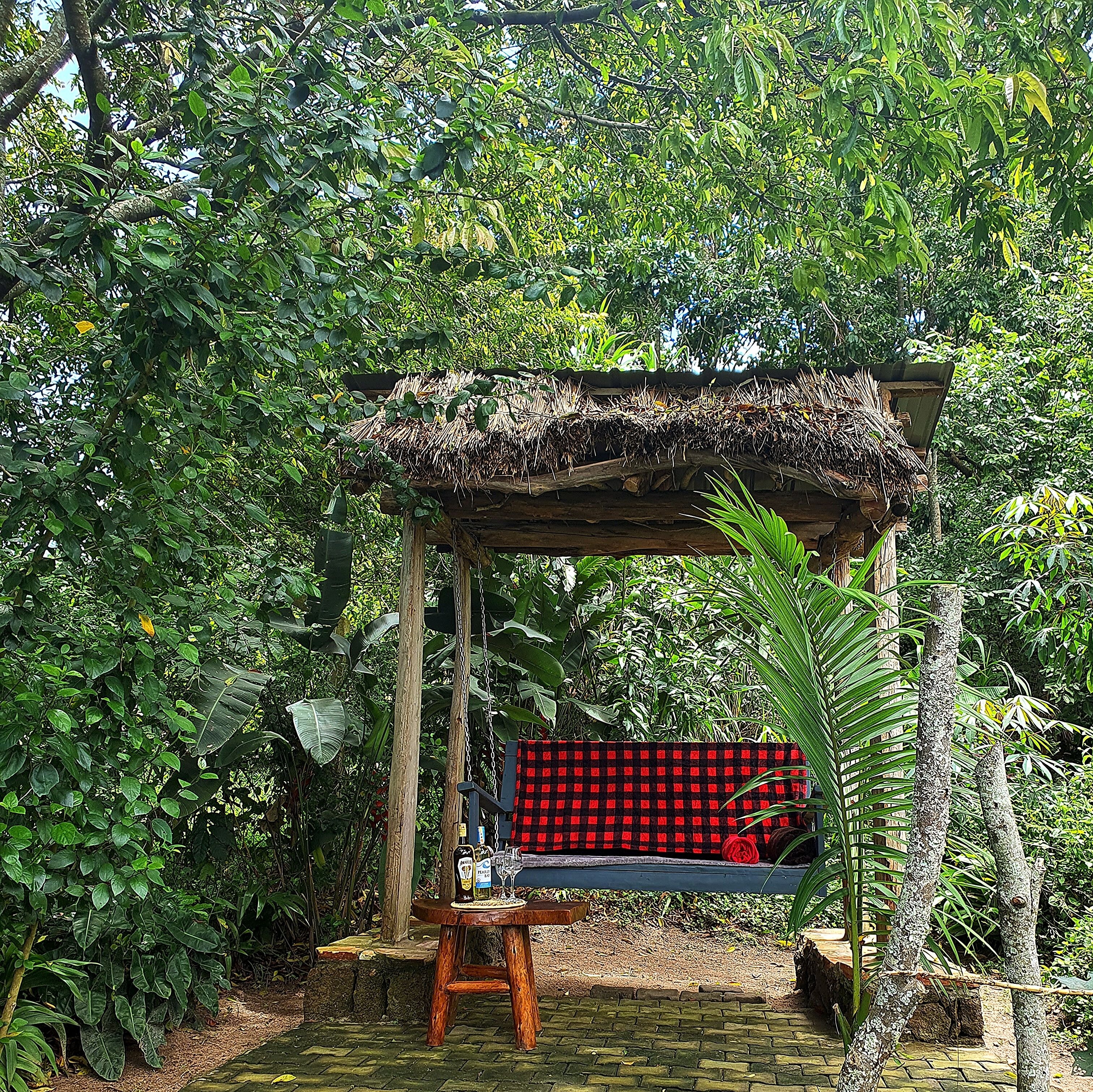 A wooden swing chair under a thatched roof structure surrounded by lush greenery.