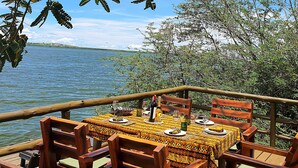 Un área de comedor al aire libre con una mesa preparada para cuatro personas, rodeada de sillas de madera con cojines verdes, con vistas a una masa de agua y una costa lejana.