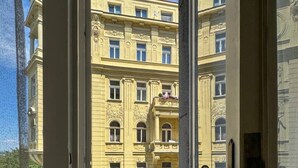 Een kamer met een deur die leidt naar een balkon, een rode stoel en een gebouw met klassieke architectuur op de achtergrond.