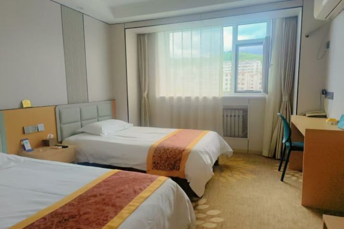 Quarto de hotel com duas camas, uma escrivaninha, uma cadeira, janela com cortinas e vista para edifícios.