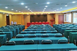 Un salón de conferencias con mesas y sillas dispuestas en filas, un escenario y una pantalla de proyección.