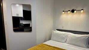 Une chambre à coucher avec un lit, un plaid jaune, une tête de lit à motifs géométriques, un miroir fixé au mur et un coin cuisine visible à travers le miroir.