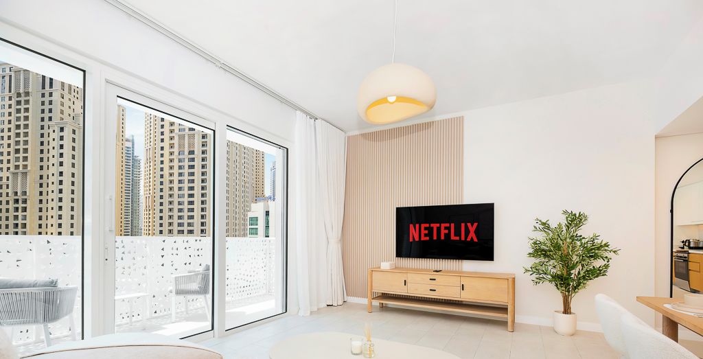 Un salon moderne doté d’un grand écran de télévision affichant le logo « NETFLIX », d’un canapé, d’une table basse et d’une vue sur de hauts immeubles à travers de grandes fenêtres.