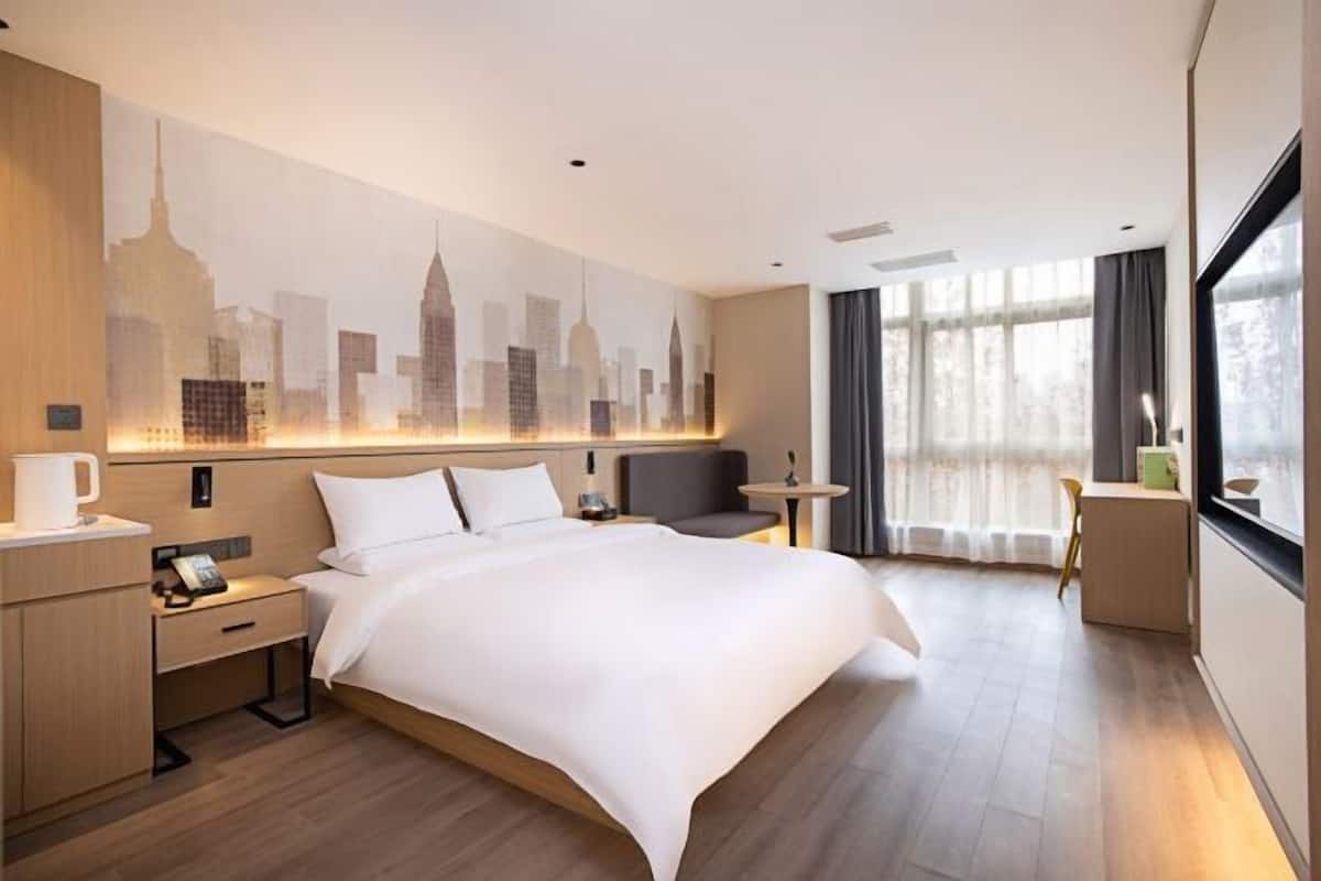 Habitación de hotel con una cama grande, una mesita de noche, un escritorio, una silla y un mural con un paisaje urbano en la pared.