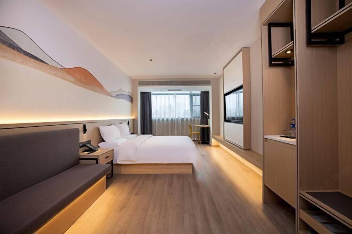 Habitación de hotel moderna con cama, mesita de noche, lámpara y vistas de la ciudad a través de amplias ventanas.