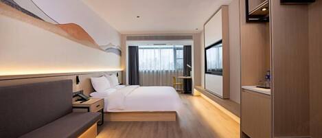 A modern hotel room with a bed, bedside table, lamp, and a view of the city through large windows.