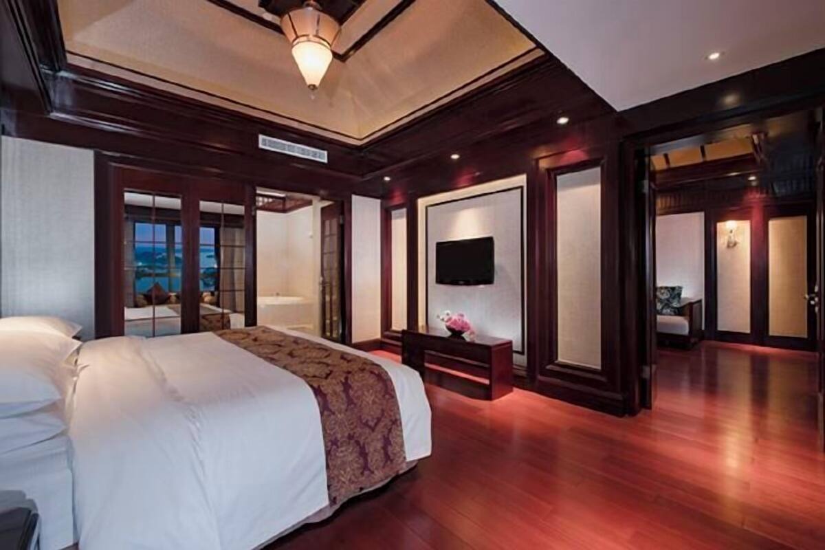 A hotel room with a large bed, a flat-screen TV mounted on the wall, and a view of the sea through a glass door.