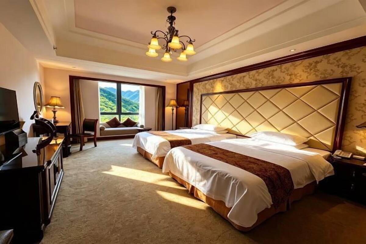 A hotel room with two beds, a chandelier, a TV, and a view of greenery outside.