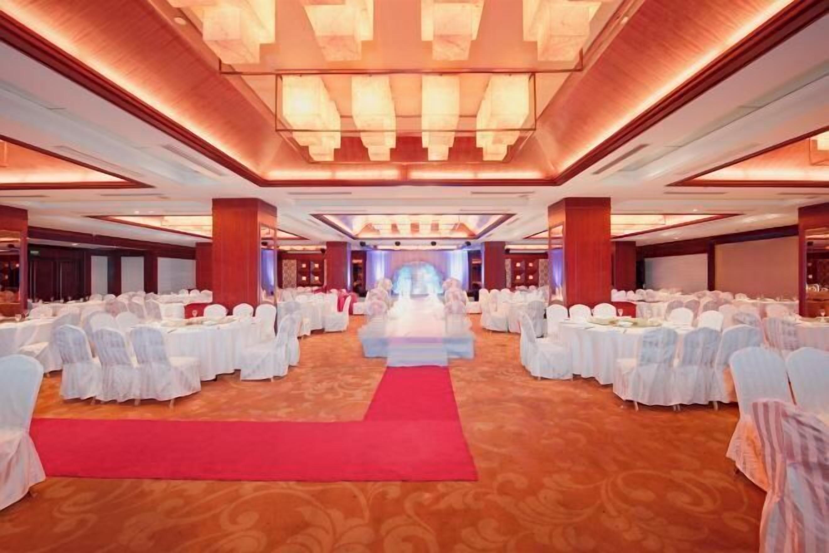 A large banquet hall with rows of round tables covered in white tablecloths and chairs, a red carpet leading to the center, and a modern chandelier design on the ceiling.