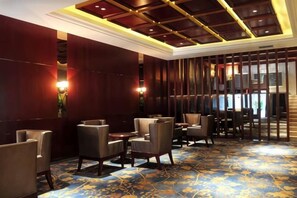 Hall d'hotel con tappeto decorato, zona salotto con sedie e tavoli, e parete divisoria in legno.