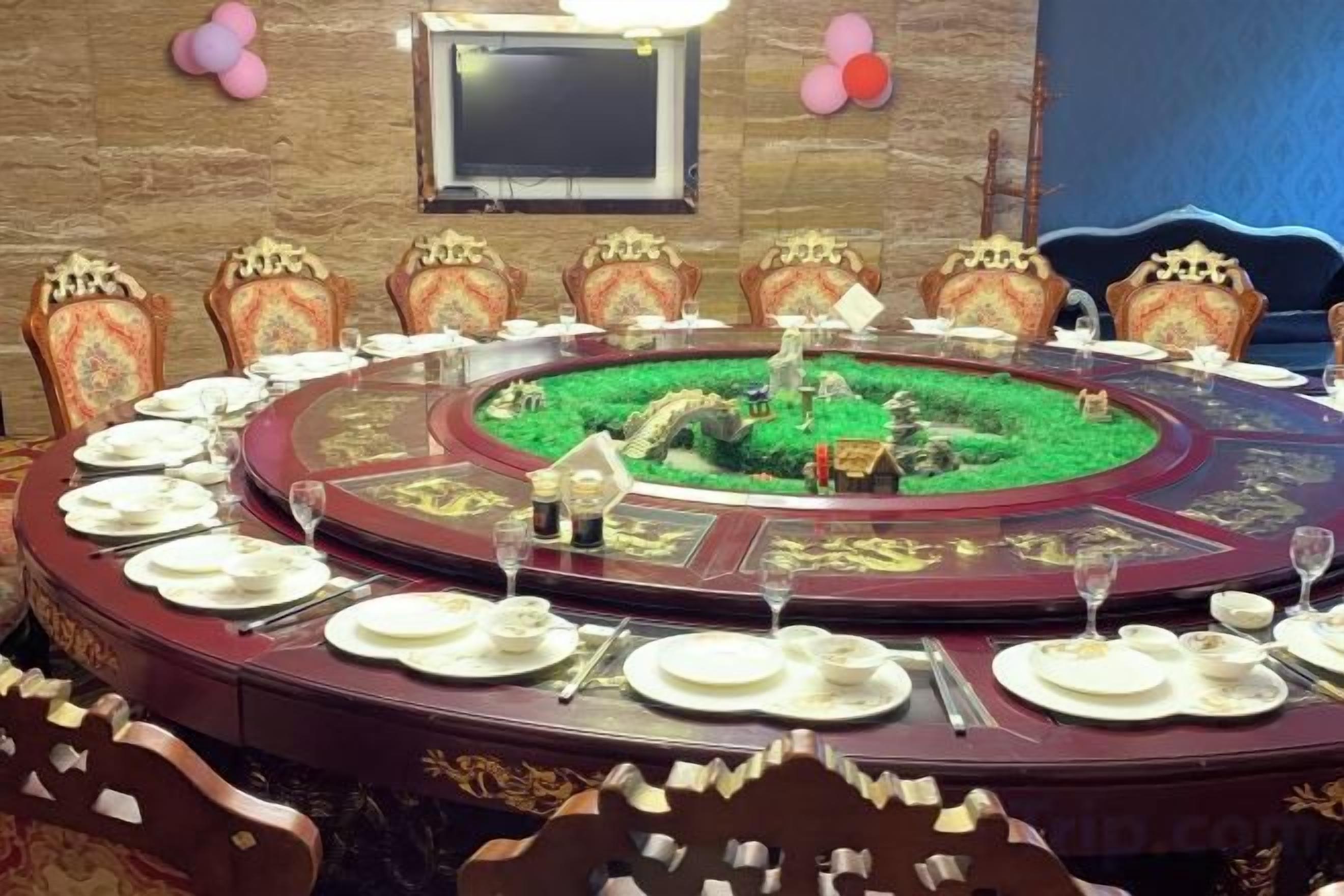 A round table with a green centerpiece, surrounded by ornate chairs, in a room with a stone wall and a television mounted above.