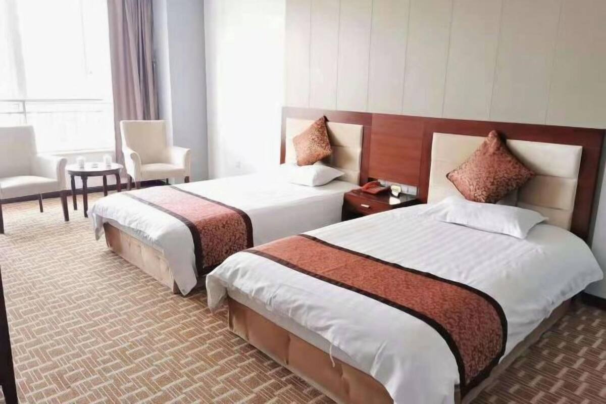 A hotel room with two beds, a small table, a chair, and a view of the outdoors.