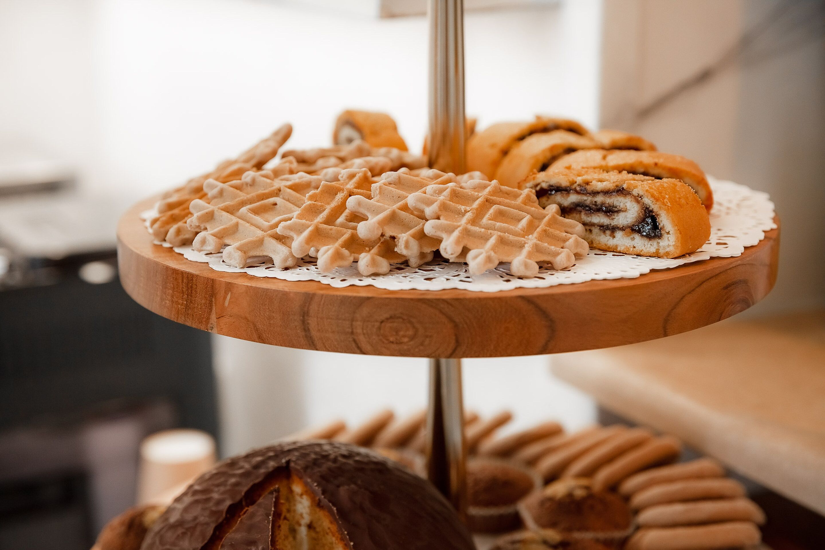 A wooden three-tiered stand with various pastries, including waffles and a cake, displayed on a lace doily.