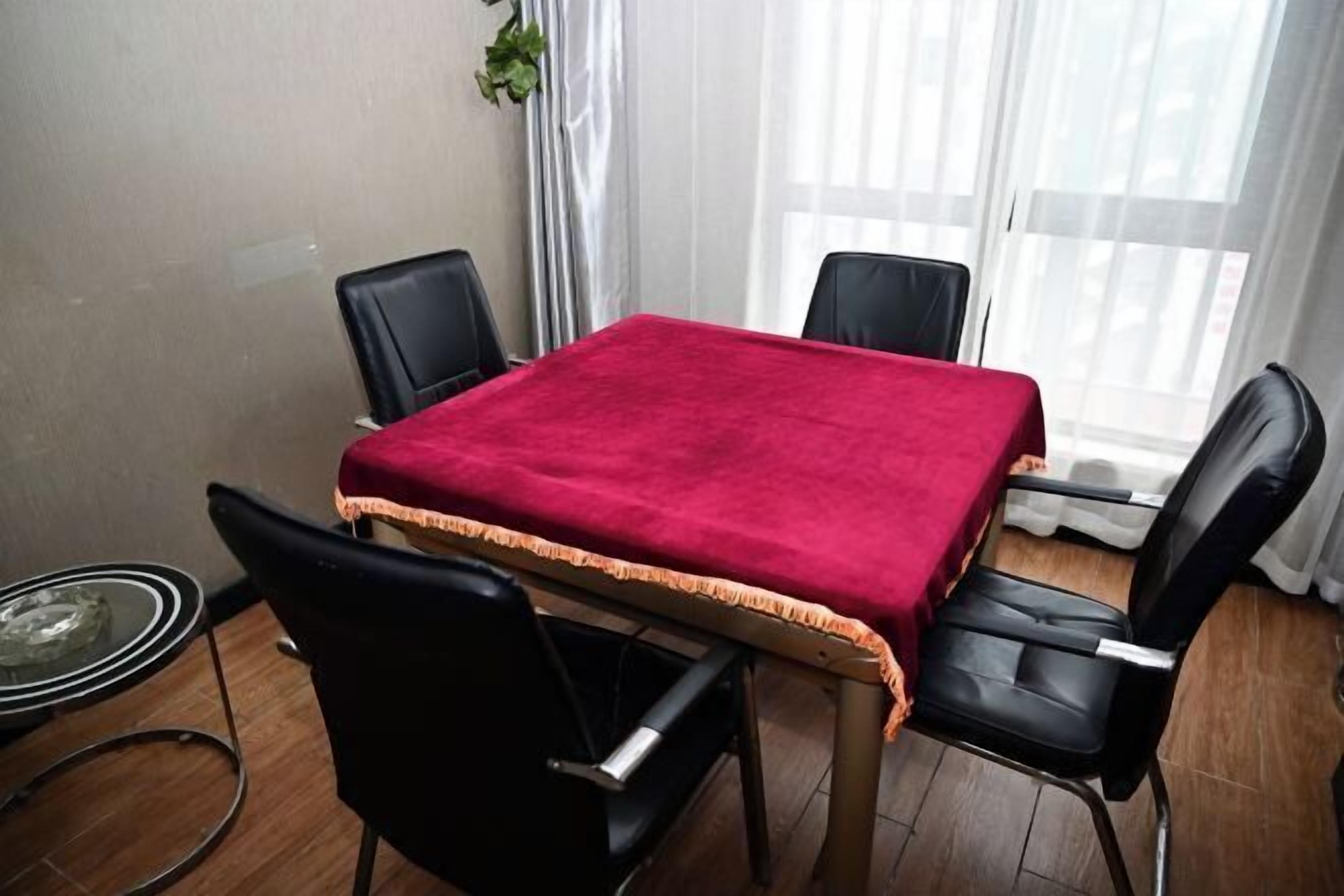 A room with a table covered by a red cloth, four black chairs, a glass table, and a window with curtains.