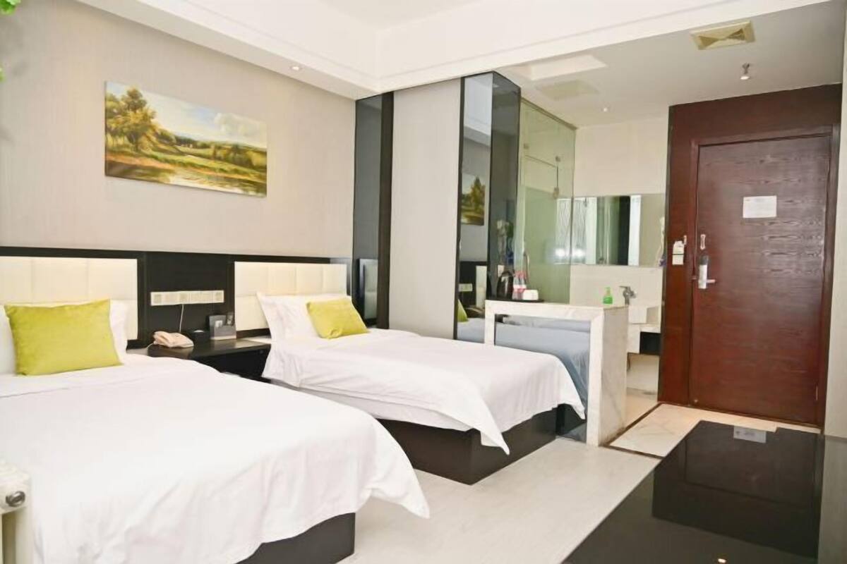 Habitación de hotel con dos camas, una mesita de noche con lámpara, un espejo, una puerta de madera y un cuadro en la pared.