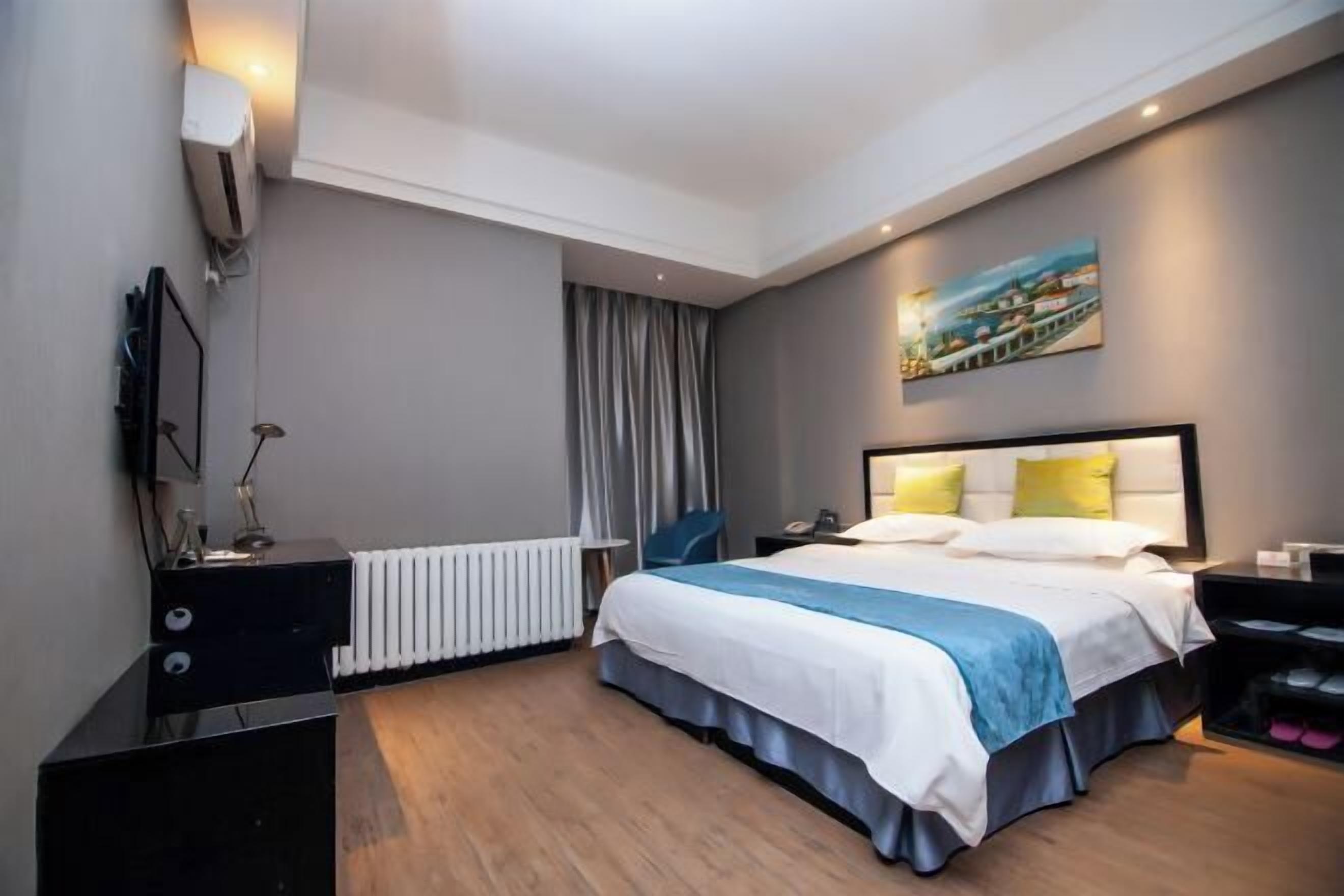 A modern hotel room with a large bed, a bedside table, a desk, a chair, a radiator, a painting on the wall, and a window with curtains.
