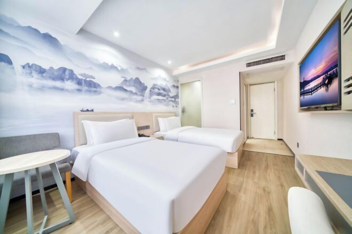 A hotel room with a large bed, a smaller bed, a desk, and a chair. There is a painting on the wall and a door leading to another room.