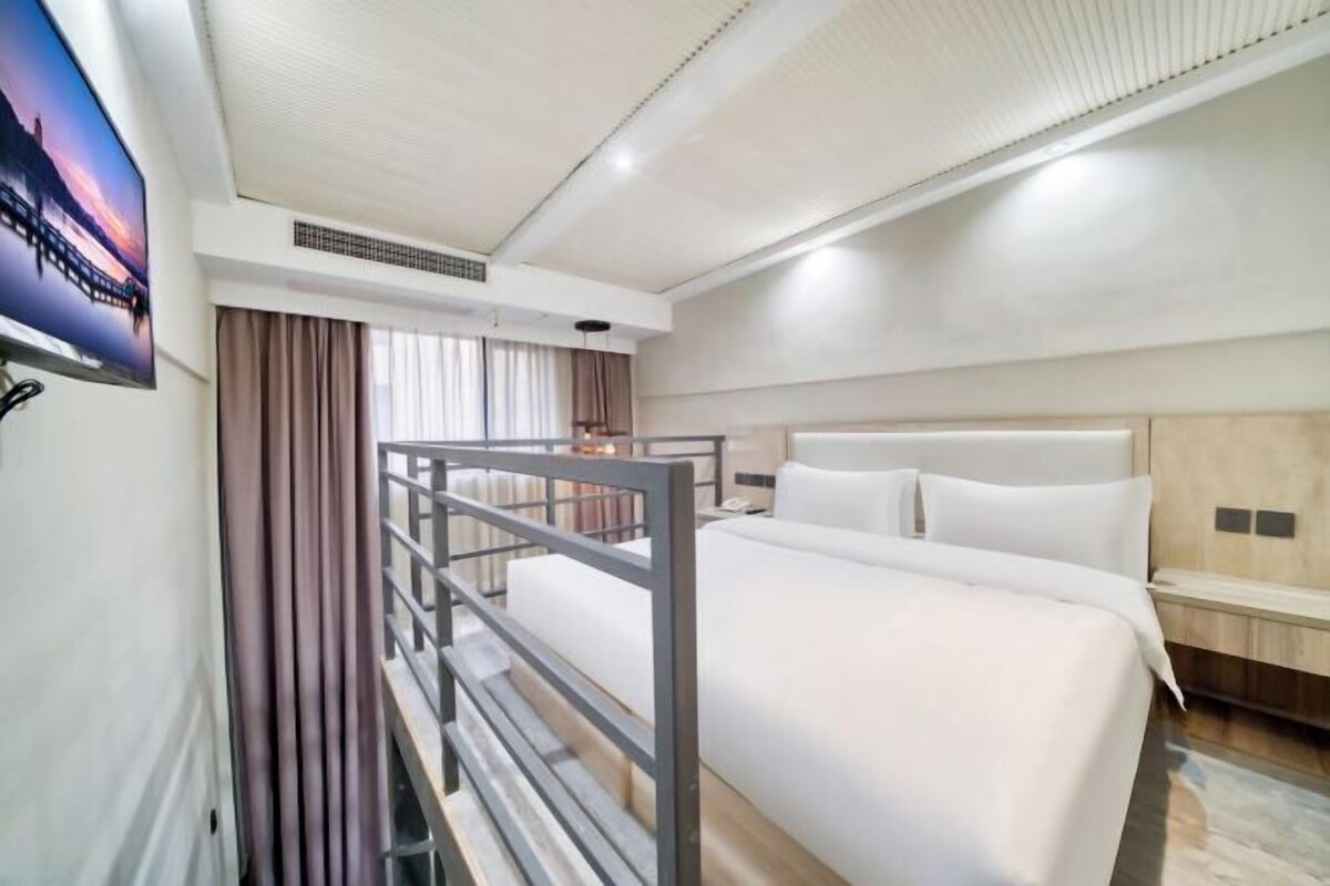 A hotel room with a bed, a TV mounted on the wall, a wooden nightstand, and a window with curtains.
