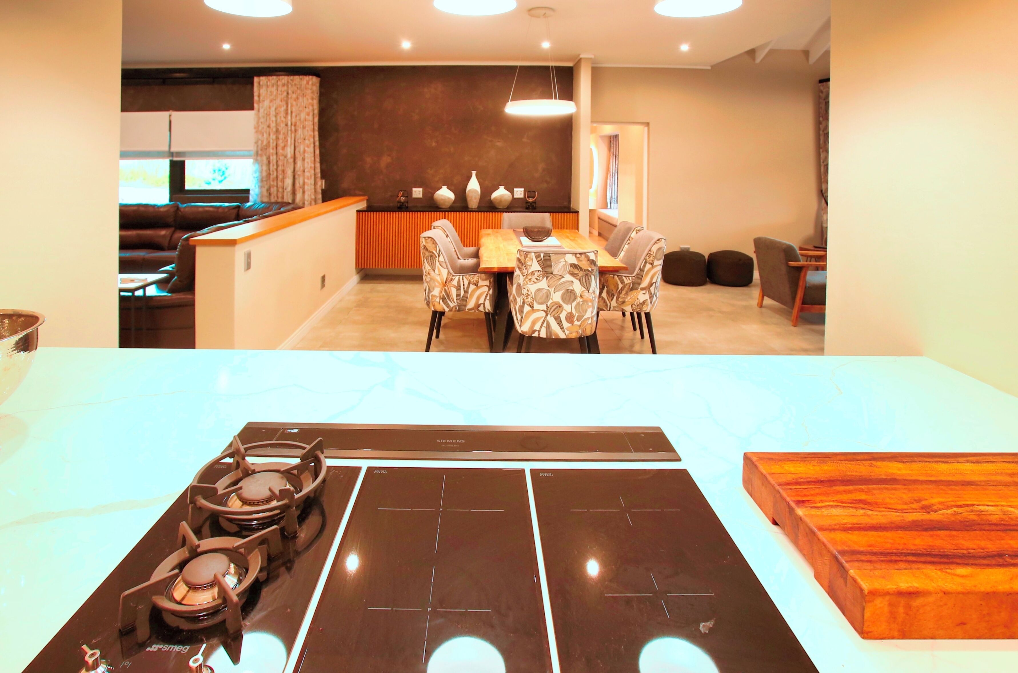 A modern kitchen with a gas stove, a dining area with a wooden table and chairs, and a living area with a sofa and a TV.