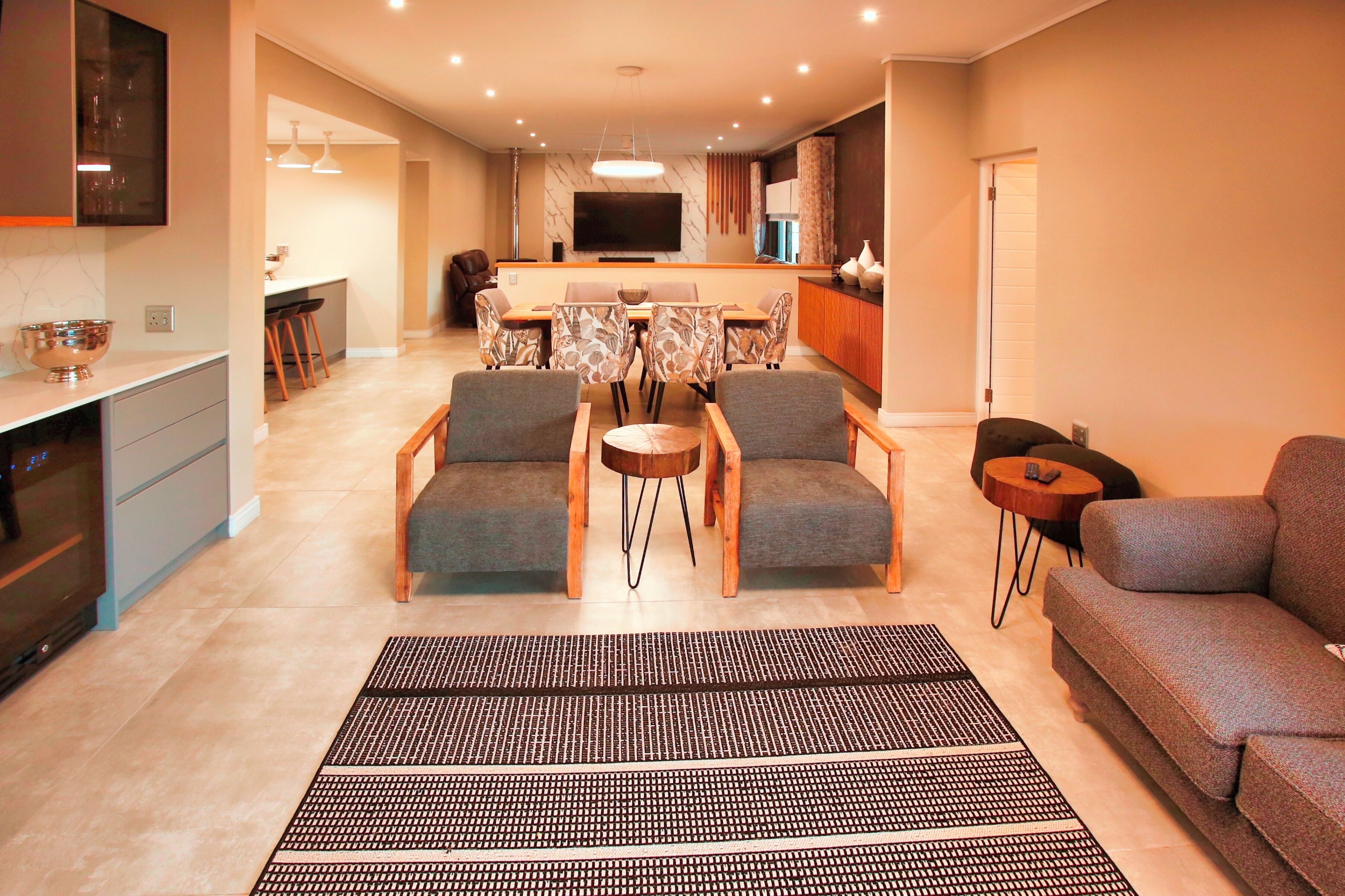 Una sala moderna con un sofá, dos sillones, una mesa de centro y un televisor. Se observa una cocina con una mesa de comedor y sillas al fondo.