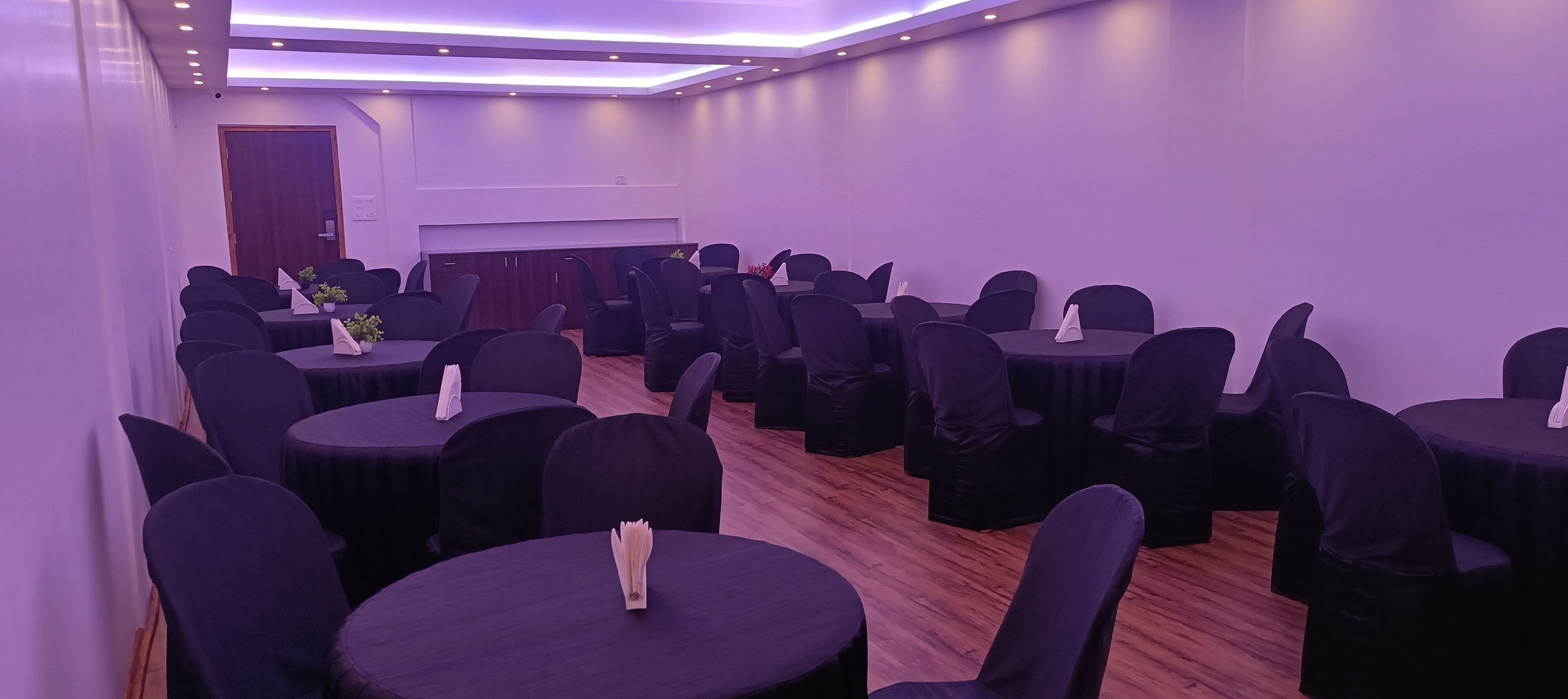 A conference room with round tables and chairs, a wooden floor, and a door in the background.
